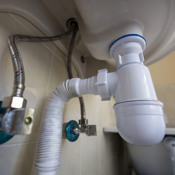 Bottom view of new white washbasin sink connected to sewer on background of bathroom light beige ceramic tiles. Professional plumbing and installation concept.